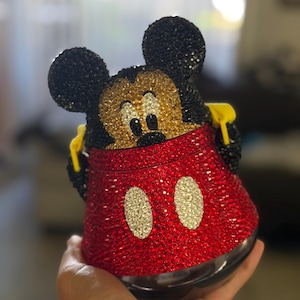 May include: A decorative container shaped like Mickey Mouse, fully covered in sparkling red, black, gold, and white rhinestones. The design features the character's iconic black ears, a gold face, and a red base with two white oval accents.