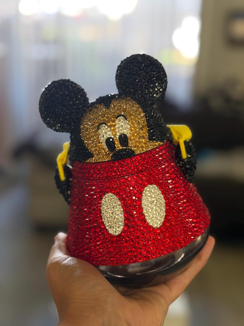May include: A decorative container shaped like Mickey Mouse, fully covered in sparkling red, black, gold, and white rhinestones. The design features the character's iconic black ears, a gold face, and a red base with two white oval accents.