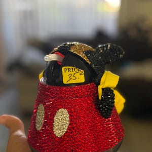 May include: A black plastic cup covered in red, gold, and black rhinestones, featuring two white oval rhinestone accents on the front, a yellow fabric bow on the side, and a clear straw, with a yellow sticker reading PRICE 35.00 attached.