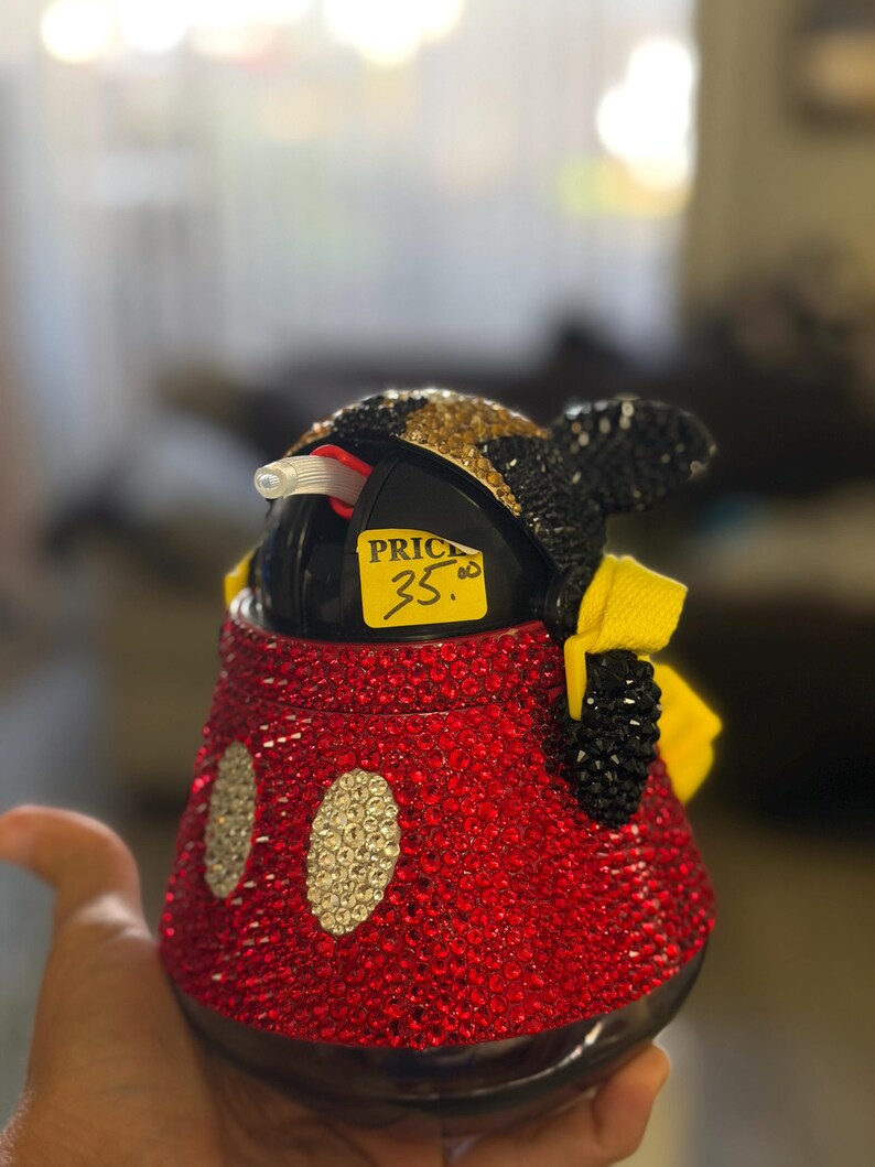 May include: A black plastic cup covered in red, gold, and black rhinestones, featuring two white oval rhinestone accents on the front, a yellow fabric bow on the side, and a clear straw, with a yellow sticker reading PRICE 35.00 attached.