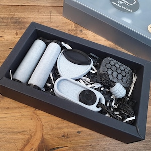 May include: A black gift box containing various grey and black accessories. The set includes two cylindrical items, a circular item, a rectangular item, and a small, round item with a black cord. The box is on a wooden surface.