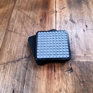 Puede incluir: Dos almohadillas adhesivas cuadradas de color negro con una superficie texturizada. Las almohadillas están diseñadas para evitar que los objetos se deslicen sobre superficies lisas.