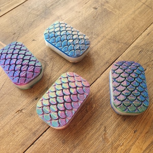 May include: Four iridescent soap bars with a mermaid scale design. The bars are rectangular and have a white base with a layer of iridescent blue, green, and purple scales on top.