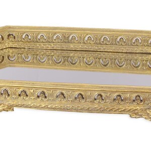 Può includere: Un vassoio da toeletta ottagonale color oro con base a specchio. Il vassoio presenta motivi ornamentali ripetuti lungo i lati e poggia su quattro piedini decorativi. Il vassoio è probabilmente in metallo.