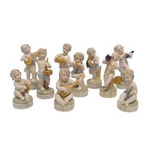 Puede incluir: Colección de once figuras de querubines de porcelana, cada una tocando un instrumento musical diferente. Los querubines están desnudos, con rasgos faciales detallados y montados sobre bases decorativas. Instrumentos: guitarra, tambor, acordeón y trompeta.
