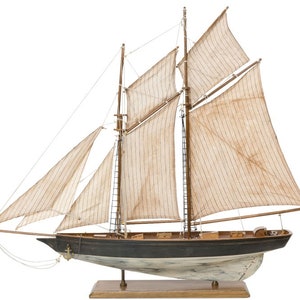 Könnte beinhalten: Ein detailliertes Modell eines Holzsegelboots mit weißen Segeln und einem schwarzen Rumpf. Das Schiff ist auf einem Holzsockel montiert.