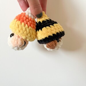 Puede incluir: Dos juguetes de peluche tejidos a crochet con caras. Uno lleva un gorro de ma&iacute;z dulce amarillo, naranja y blanco. El otro lleva un gorro a rayas negro y amarillo.