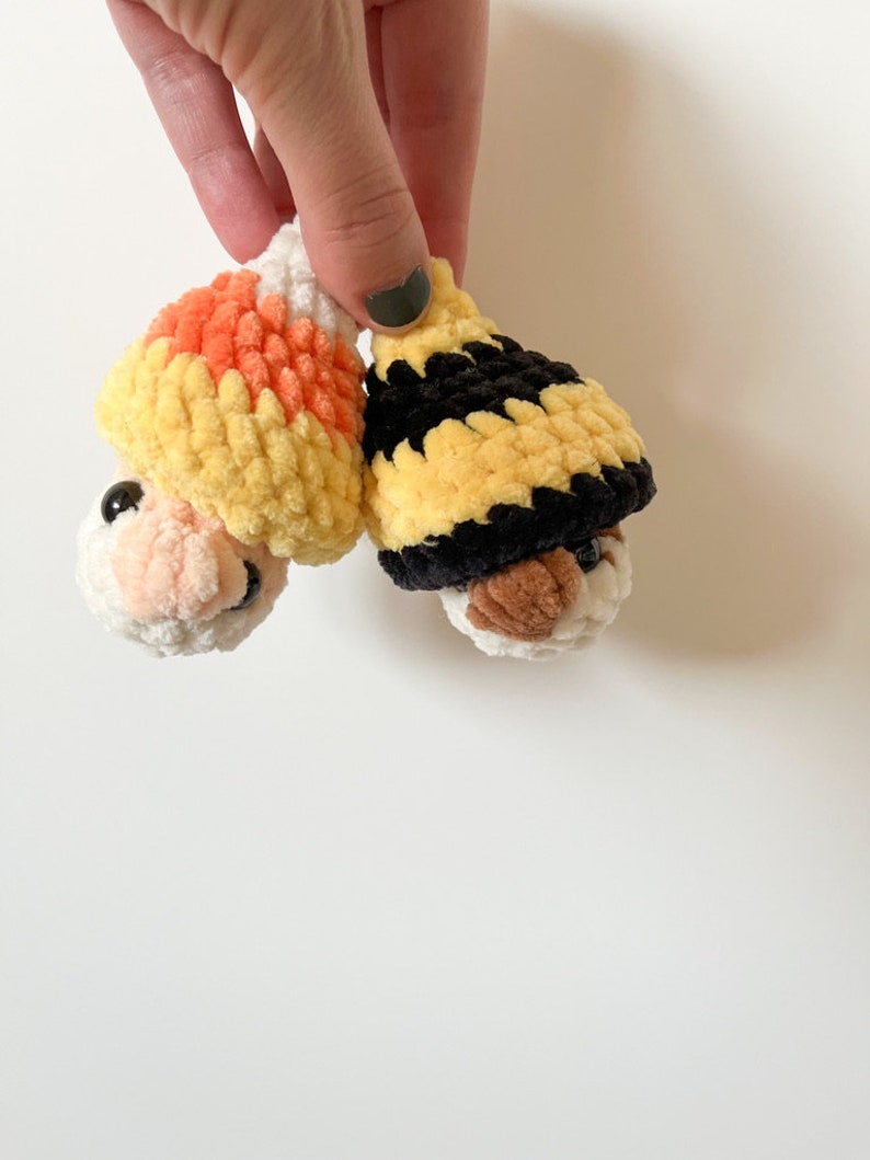 Puede incluir: Dos juguetes de peluche tejidos a crochet con caras. Uno lleva un gorro de ma&iacute;z dulce amarillo, naranja y blanco. El otro lleva un gorro a rayas negro y amarillo.