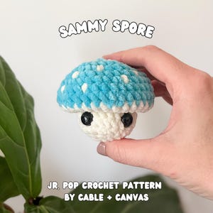 May include: A handmade crochet mushroom toy. The mushroom has a light blue cap with white spots, a white stem, and black eyes. The text "SAMMY SPORE" is above the mushroom, and "JR. POP CROCHET PATTERN BY CABLE + CANVAS" is below.
