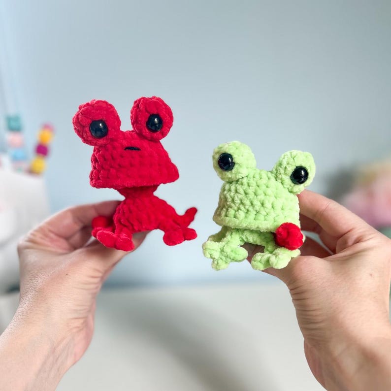 May include: Two crocheted frog toys. One is bright red, the other is light green. Both have large, black eyes and small red accents. The frogs are handmade and have a soft, textured appearance. The toys are small and appear to be held in someone's hands.