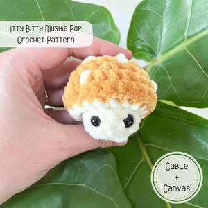 Puede incluir: Un juguete de amigurumi de hongo tejido a crochet con un cuerpo blanco y una tapa marr&oacute;n peluda. El juguete tiene ojos negros y se sostiene en una mano. El texto "itty Bitty Mushie Pop Crochet Pattern" es visible en la imagen. El texto "Cable + Canvas" tambi&eacute;n es visible en la imagen.