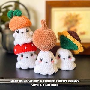 May include: Crocheted ghost figures with various hats, including an orange mushroom, a red mushroom with white spots, and a brown acorn. The ghosts are white with black eyes and pink cheeks. The text at the bottom reads "MADE USING WEIGHT 6 PREMIER PARFAIT CHUNKY WITH A 4 MM HOOK."