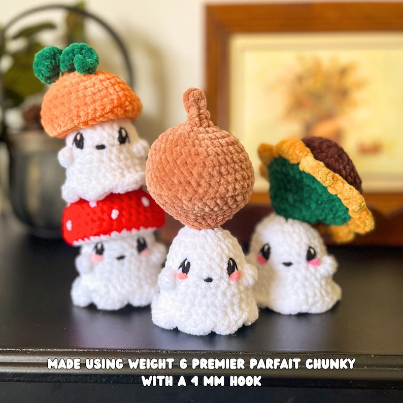 May include: Crocheted ghost figures with various hats, including an orange mushroom, a red mushroom with white spots, and a brown acorn. The ghosts are white with black eyes and pink cheeks. The text at the bottom reads "MADE USING WEIGHT 6 PREMIER PARFAIT CHUNKY WITH A 4 MM HOOK."
