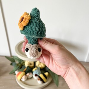 Puede incluir: Un peque&ntilde;o gnomo de crochet que lleva un sombrero verde con un girasol amarillo en la parte superior. El gnomo sostiene una peque&ntilde;a calabaza en su mano.