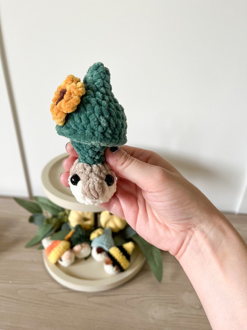 Puede incluir: Un peque&ntilde;o gnomo de crochet que lleva un sombrero verde con un girasol amarillo en la parte superior. El gnomo sostiene una peque&ntilde;a calabaza en su mano.