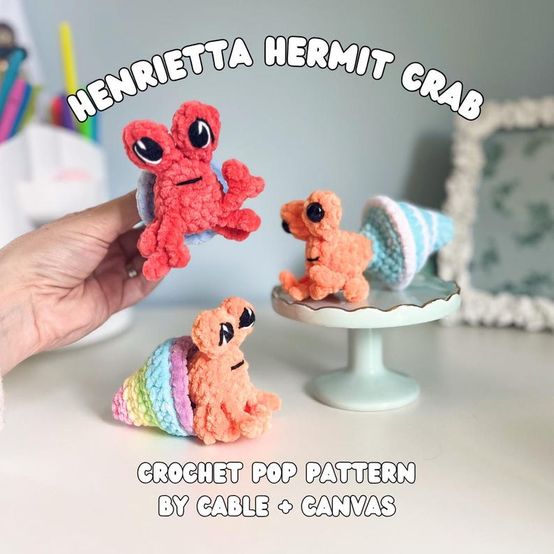 May include: Three crochet hermit crab toys in shades of orange and red. One crab is held in a hand, while the others sit on a small cake stand. The crabs have black eyes and are accompanied by the text "HENRIETTA HERMIT CRAB" and "CROCHET POP PATTERN BY CABLE + CANVAS."