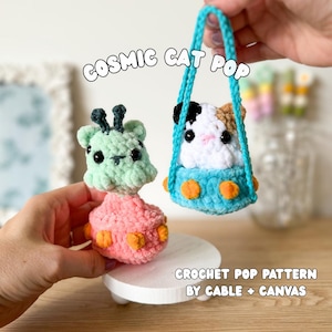 May include: Two crocheted plush toys featuring a light green creature with black antennae and a white, black, and tan cat inside a blue UFO-shaped vessel with orange accents. The text reads COSMIC CAT POP and CROCHET POP PATTERN BY CABLE + CANVAS.
