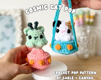 May include: Two crocheted plush toys featuring a light green creature with black antennae and a white, black, and tan cat inside a blue UFO-shaped vessel with orange accents. The text reads COSMIC CAT POP and CROCHET POP PATTERN BY CABLE + CANVAS.
