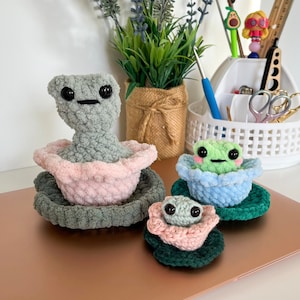 May include: Three crocheted frog plushies, each with a different colored base. The largest frog is gray with black eyes and a pink base. The middle frog is green with black eyes and a light blue base. The smallest frog is gray with black eyes and a pink base.
