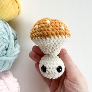 May include: A handmade crochet mushroom toy with a brown cap dotted with white spots and a white stem. The mushroom has large black safety eyes. Soft yarn in yellow, blue, and pink are in the background.
