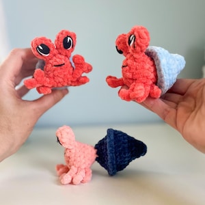 May include: Three handmade crochet hermit crabs in shades of red, pink, and blue. The crabs have detailed features, including black eyes and small claws. The crab shells are blue and the bodies are red or pink.