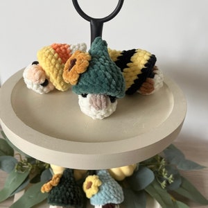 Puede incluir: Una bandeja de madera de dos niveles con un asa de metal negro. El nivel superior tiene tres gnomos de crochet que llevan diferentes sombreros: un sombrero de ma&iacute;z dulce amarillo y naranja, un sombrero verde con una flor amarilla y un sombrero a rayas negro y amarillo. El nivel inferior tiene dos gnomos m&aacute;s que llevan sombreros verdes y azules con flores amarillas.