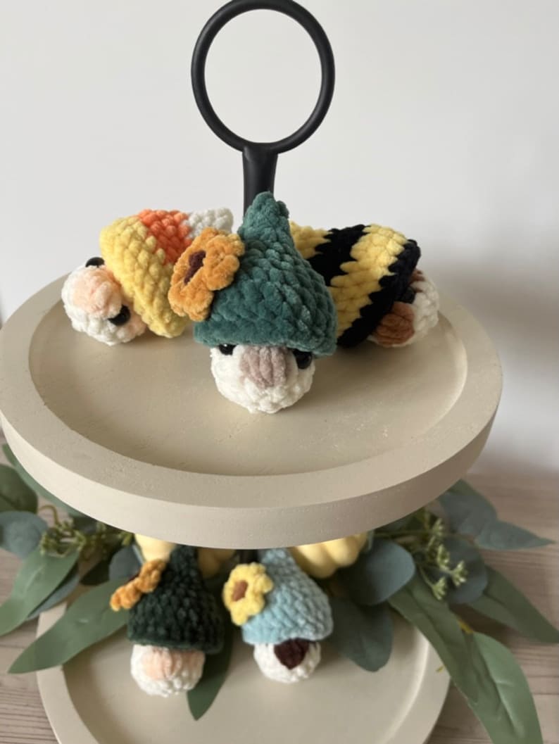 Puede incluir: Una bandeja de madera de dos niveles con un asa de metal negro. El nivel superior tiene tres gnomos de crochet que llevan diferentes sombreros: un sombrero de ma&iacute;z dulce amarillo y naranja, un sombrero verde con una flor amarilla y un sombrero a rayas negro y amarillo. El nivel inferior tiene dos gnomos m&aacute;s que llevan sombreros verdes y azules con flores amarillas.