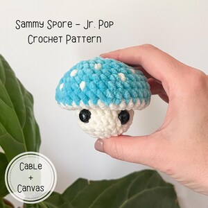 Puede incluir: Un juguete de amigurumi de hongo tejido a crochet con un gorro azul y blanco y un tallo blanco. El hongo tiene ojos negros y est&aacute; siendo sostenido por una mano.