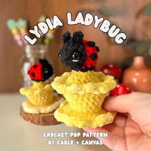 Op de afbeelding: Een handgemaakte gehaakte lieveheersbeestjesfiguur met een zwarte kop en rood gestippelde vleugels, bovenop een gele gerimpelde basis. De tekst "LYDIA LADYBUG" en "CROCHET POP PATTERN BY CABLE + CANVAS" zijn zichtbaar.