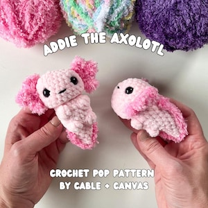 May include: Two handmade crochet axolotl toys in light pink, held by hands. The axolotls have black eyes and pink frills. The text "Addie the Axolotl" and "Crochet Pop Pattern by Cable + Canvas" are visible.
