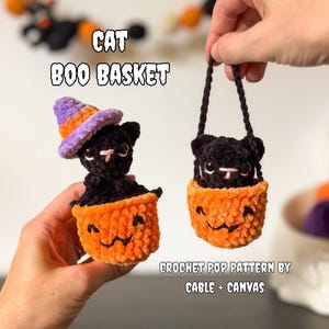 May include: Two small black crochet cats in orange pumpkin-shaped baskets. One cat wears a purple and orange witch hat. The text reads CAT BOO BASKET and CROCHET POP PATTERN BY CABLE + CANVAS. The baskets feature embroidered black faces.