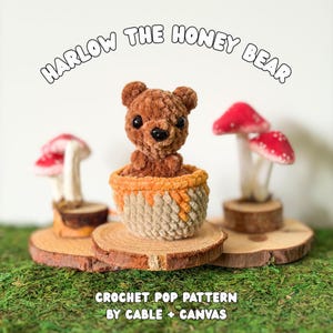 May include: A brown crochet bear sitting in a tan and orange honey pot, flanked by two red and white mushroom figures. The text reads HARLOW THE HONEY BEAR and CROCHET POP PATTERN BY CABLE + CANVAS. The items are displayed on wooden slices.