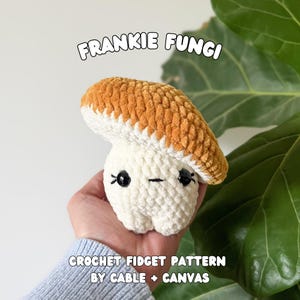 May include: A handmade crochet mushroom toy with a white stem and a textured orange cap. The mushroom has black eyes and a small smile. Text on the image reads "FRANKIE FUNGI" and "CROCHET FIDGET PATTERN BY CABLE + CANVAS."