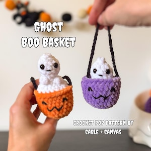 Op de afbeelding: Twee kleine gehaakte spookfiguren in miniatuurmandjes. De ene mand is oranje en de andere is paars, beide met zwarte geborduurde gezichten. De spoken hebben lichamen van wit garen en zwarte veiligheidsoogjes. Tekst leest GHOST BOO BASKET.