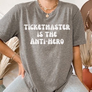 May include: A gray t-shirt with the text "Ticketmaster is the anti-hero" in white, retro-style lettering.