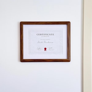Peut inclure: Un certificat d'achèvement dans un cadre en bois brun foncé avec un passe-partout blanc. Le document porte le texte "CERTIFICATE OF COMPLETION", "This Certificate is presented to", "Jack Sanders", ainsi que deux signatures et un sceau rouge.