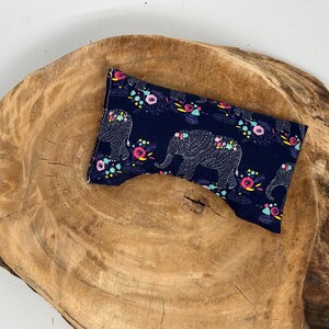 May include: A navy blue eye pillow with a repeating pattern of white elephants and colorful flowers. The pillow is rectangular with a curved indentation. It rests on a rustic, wooden surface.