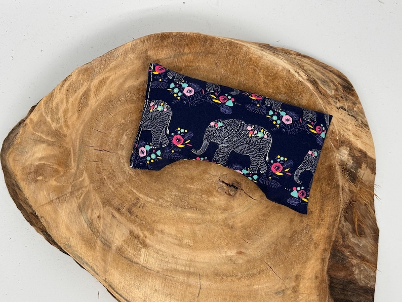 May include: A navy blue eye pillow with a repeating pattern of white elephants and colorful flowers. The pillow is rectangular with a curved indentation. It rests on a rustic, wooden surface.