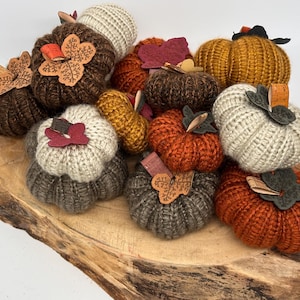 May include: A collection of handmade, knitted pumpkins in various colors, including brown, orange, and cream. Each pumpkin has a decorative stem and leaf accents, perfect for fall decor. The pumpkins are arranged on a wooden surface.