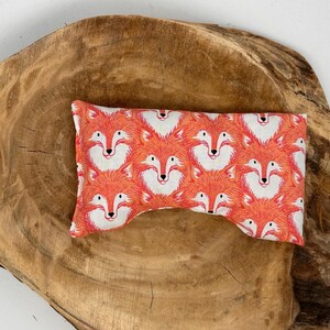 May include: A rectangular eye pillow with a repeating pattern of orange foxes with white faces. The pillow is placed on a large, round, wooden surface with a natural wood grain.
