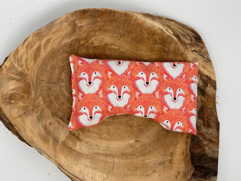 May include: A rectangular eye pillow with a repeating pattern of orange foxes with white faces. The pillow is placed on a large, round, wooden surface with a natural wood grain.