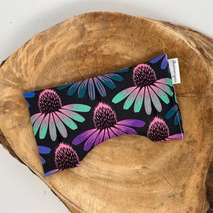 May include: A black eye pillow with a floral pattern featuring pink, purple, and teal flowers. The pillow is rectangular and rests on a wooden surface. A small white tag with text is attached to the pillow.