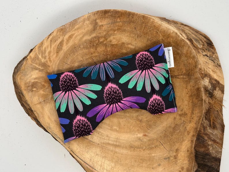 May include: A black eye pillow with a floral pattern featuring pink, purple, and teal flowers. The pillow is rectangular and rests on a wooden surface. A small white tag with text is attached to the pillow.