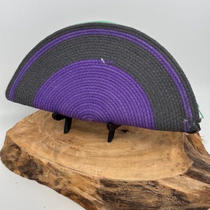 May include: A half-circle shaped, hand-woven rope basket in shades of gray, purple, and green. The basket is displayed on a wooden base with black metal supports. The rope is tightly coiled, creating a textured surface. The basket is approximately 12 inches wide.