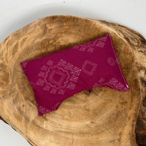 May include: A magenta-colored rectangular pillow with a white geometric pattern. The pillow is placed on a large, textured, wooden surface with a natural wood grain.
