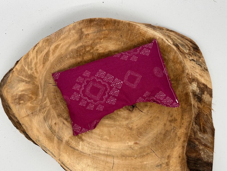 May include: A magenta-colored rectangular pillow with a white geometric pattern. The pillow is placed on a large, textured, wooden surface with a natural wood grain.