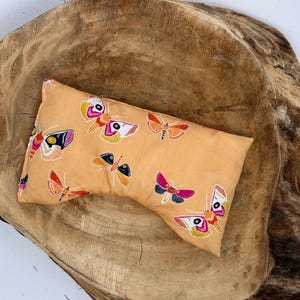 May include: A rectangular, peach-colored eye pillow featuring a repeating pattern of colorful moths. The moths have intricate wing designs in shades of pink, orange, yellow, and black. The pillow is resting on a large, textured, wooden surface.