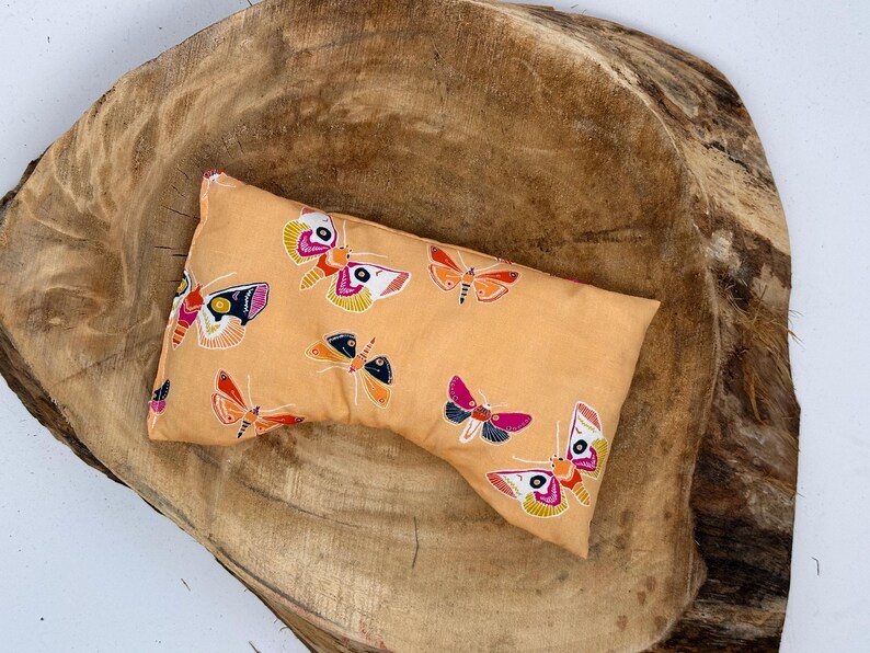 May include: A rectangular, peach-colored eye pillow featuring a repeating pattern of colorful moths. The moths have intricate wing designs in shades of pink, orange, yellow, and black. The pillow is resting on a large, textured, wooden surface.