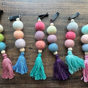May include: Six colourful felt ball car seat chain clips with tassels. Each chain has five felt balls in different colours, including pink, purple, blue, green, and grey. The chains are attached to black clips.