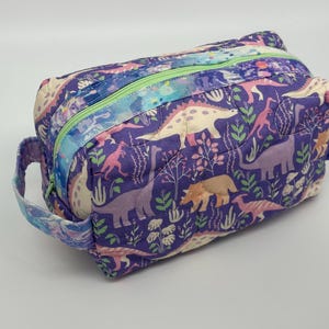 Puede incluir: Bolsa de cosméticos rectangular acolchada con estampado de dinosaurios en tonos morado, rosa y crema. La bolsa tiene una cremallera verde claro y asas a juego. Mide aproximadamente 23 cm de largo y 13 cm de alto.